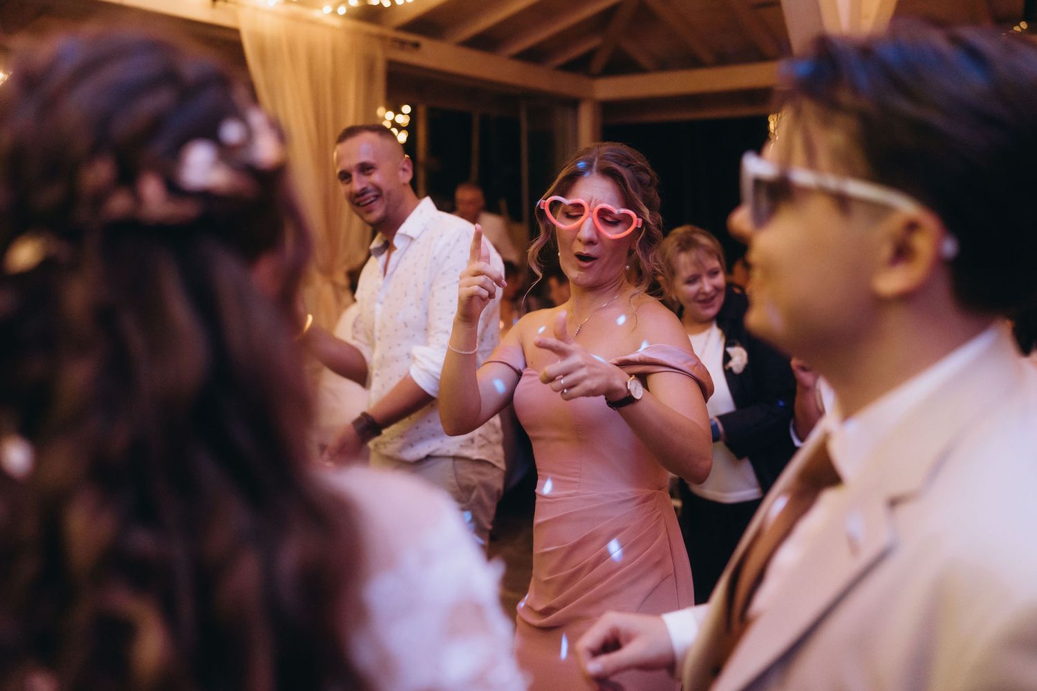 Guests dancing at wedding reception, woman in pink dress wearing heart-shaped sunglasses, festive atmosphere.