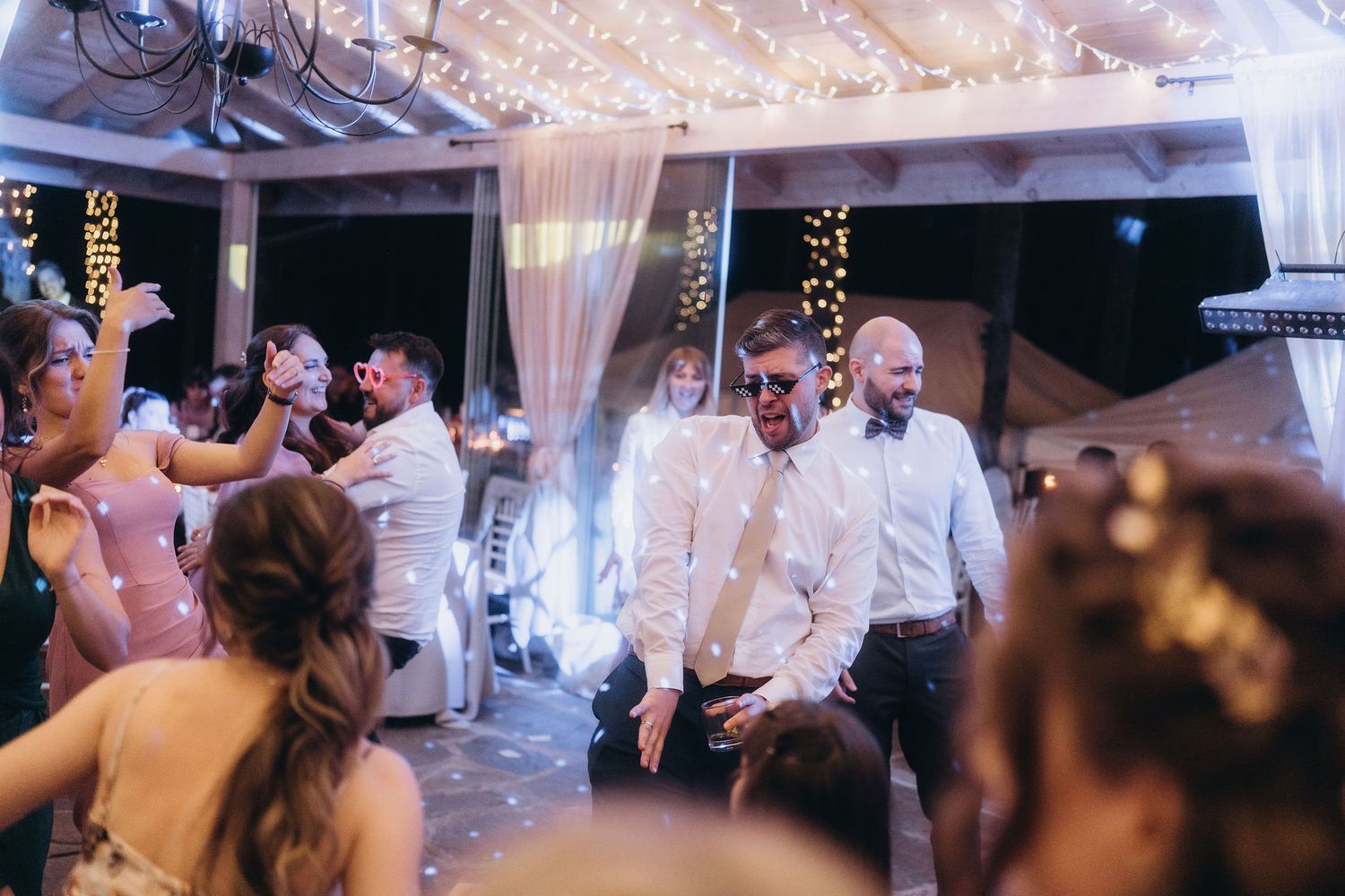 Two grooms dancing joyfully surrounded by cheering wedding guests under fairy lights.