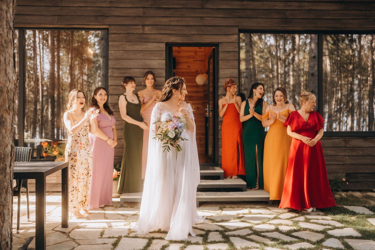 Bride in white gown holds bouquet while colorful bridesmaids applaud outside a rustic wooden cabin.
