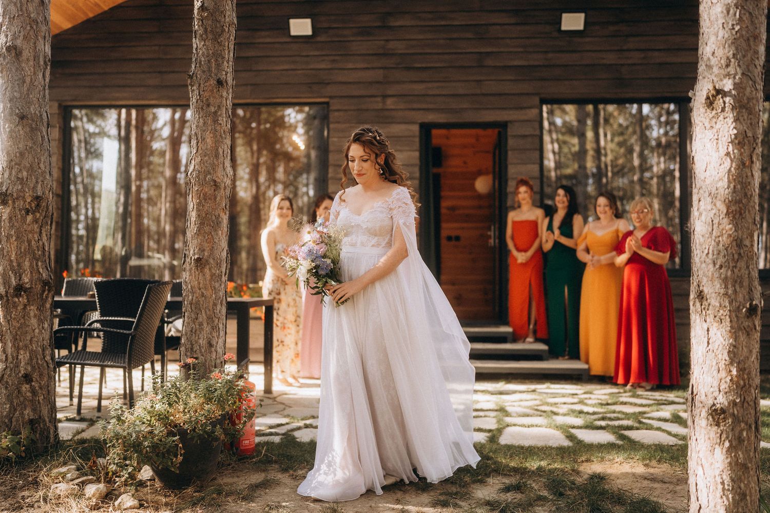 Bride in white gown holds bouquet outdoors near log cabin, colorful bridesmaids in rainbow dresses stand in background.