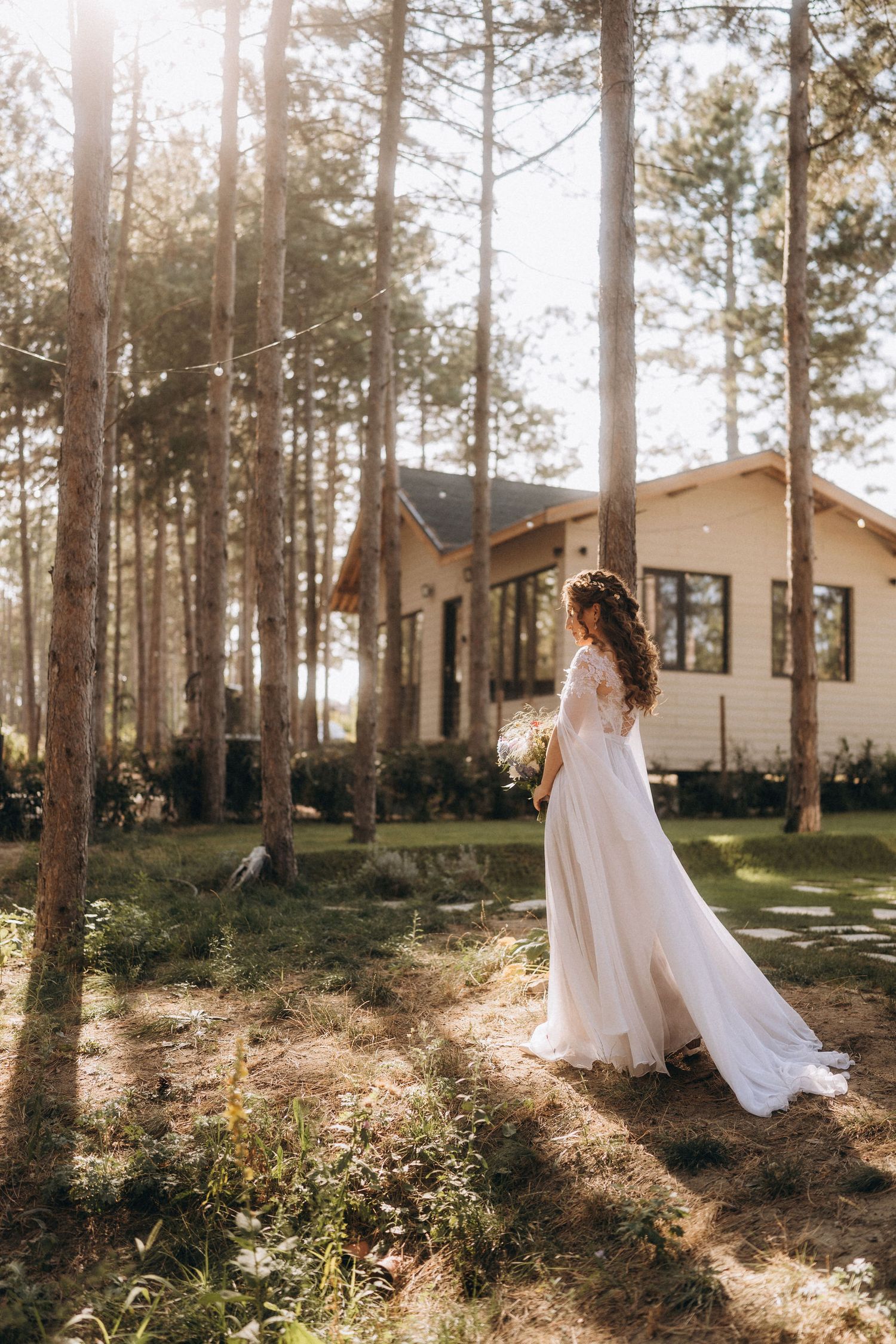 Bride in white lace gown stands among tall pine trees near a wooden cabin, bathed in golden sunlight.