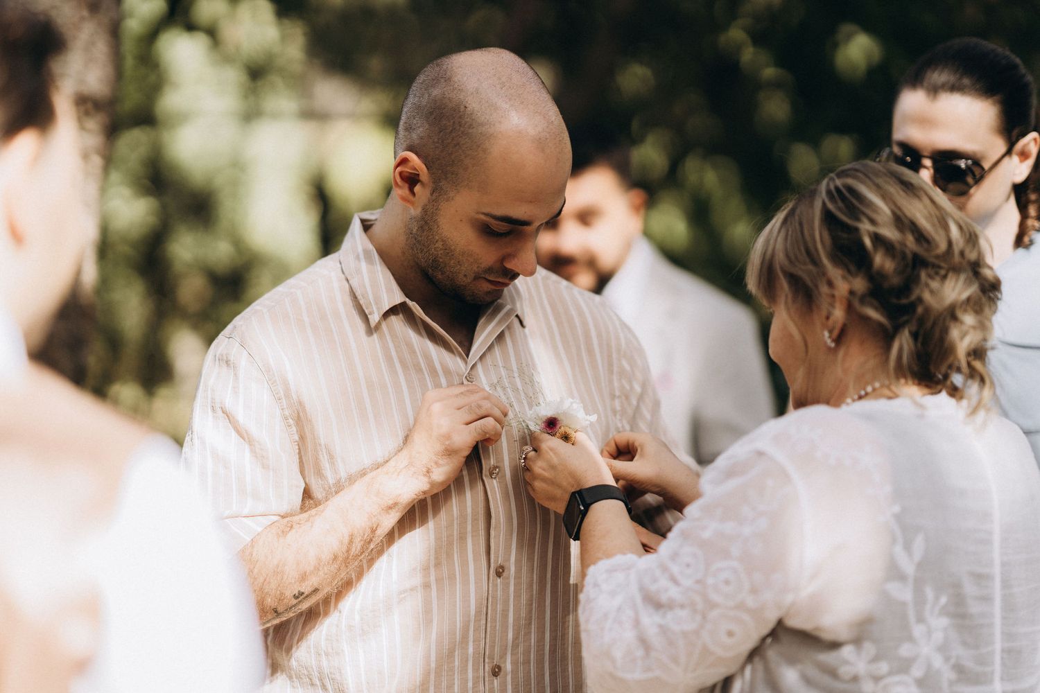 A bald groom in a striped shirt gets a boutonniere pinned by a woman at an outdoor wedding ceremony.