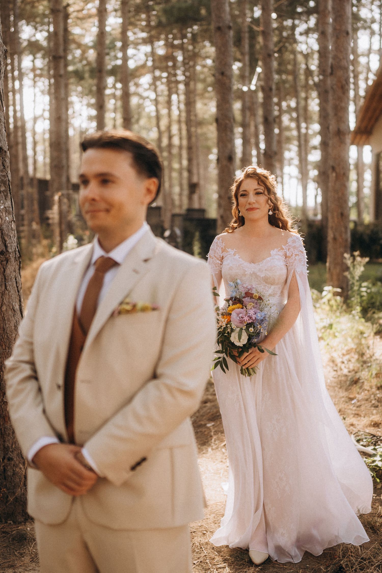 Elegant bride in lace gown holding bouquet approaches groom in beige suit in pine forest.