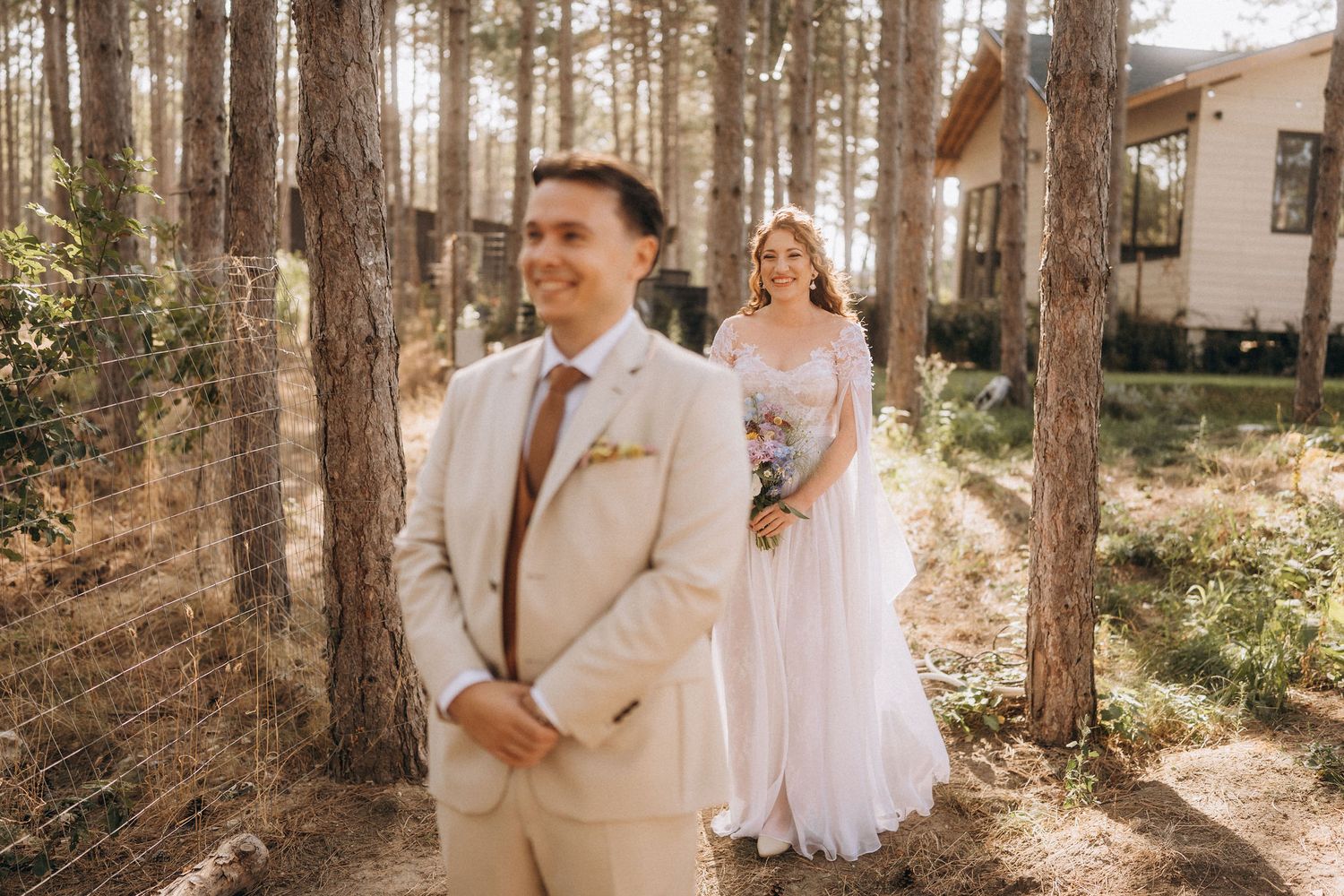 Groom in beige suit smiles as bride in white gown approaches through sunlit pine forest at outdoor wedding.
