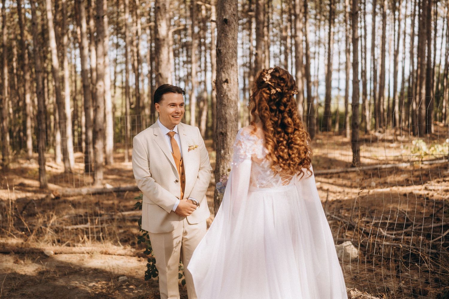 Groom in beige suit smiles at bride in white gown holding hands in a sunlit pine forest wedding.