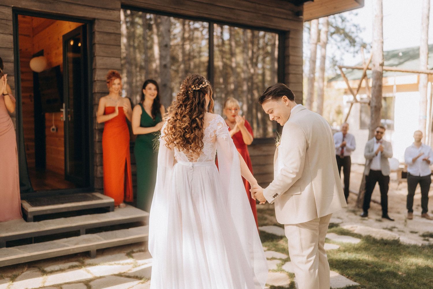 Bride and groom holding hands outdoors at wedding ceremony with guests watching near a wooden building.
