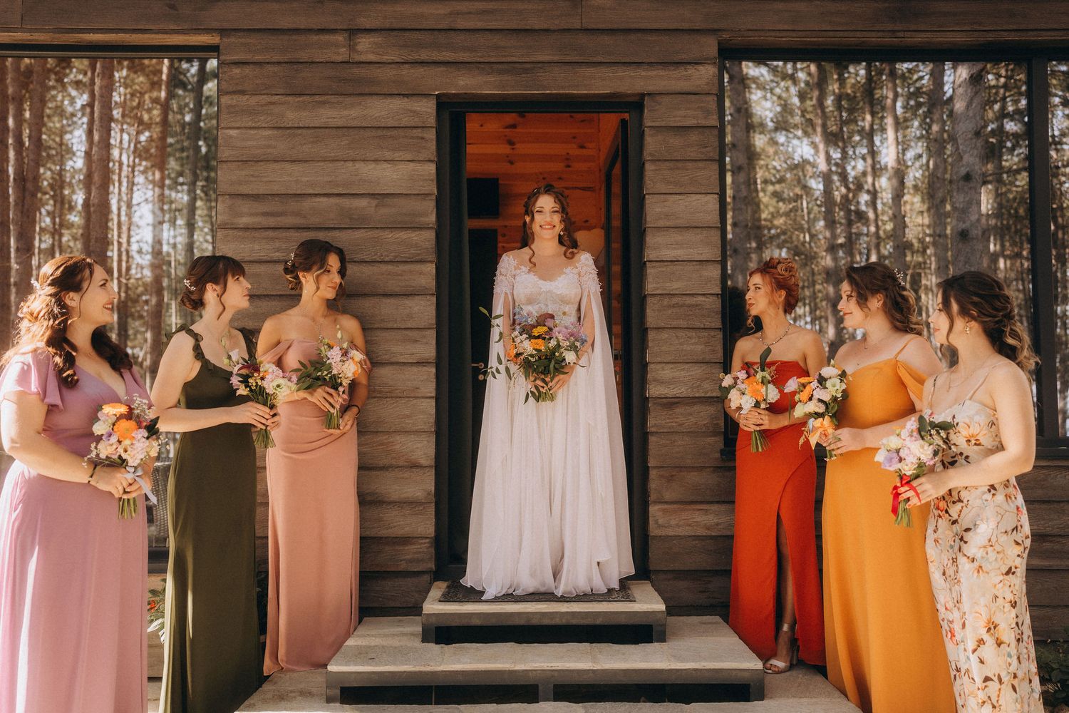 Bride in white lace gown stands on cabin steps surrounded by bridesmaids in colorful mismatched dresses.