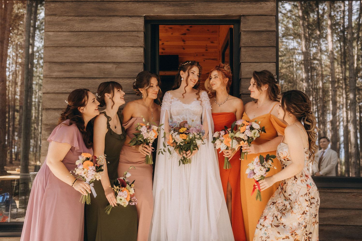 Bride and five bridesmaids in colorful dresses laugh together outside a rustic wooden cabin in the forest.