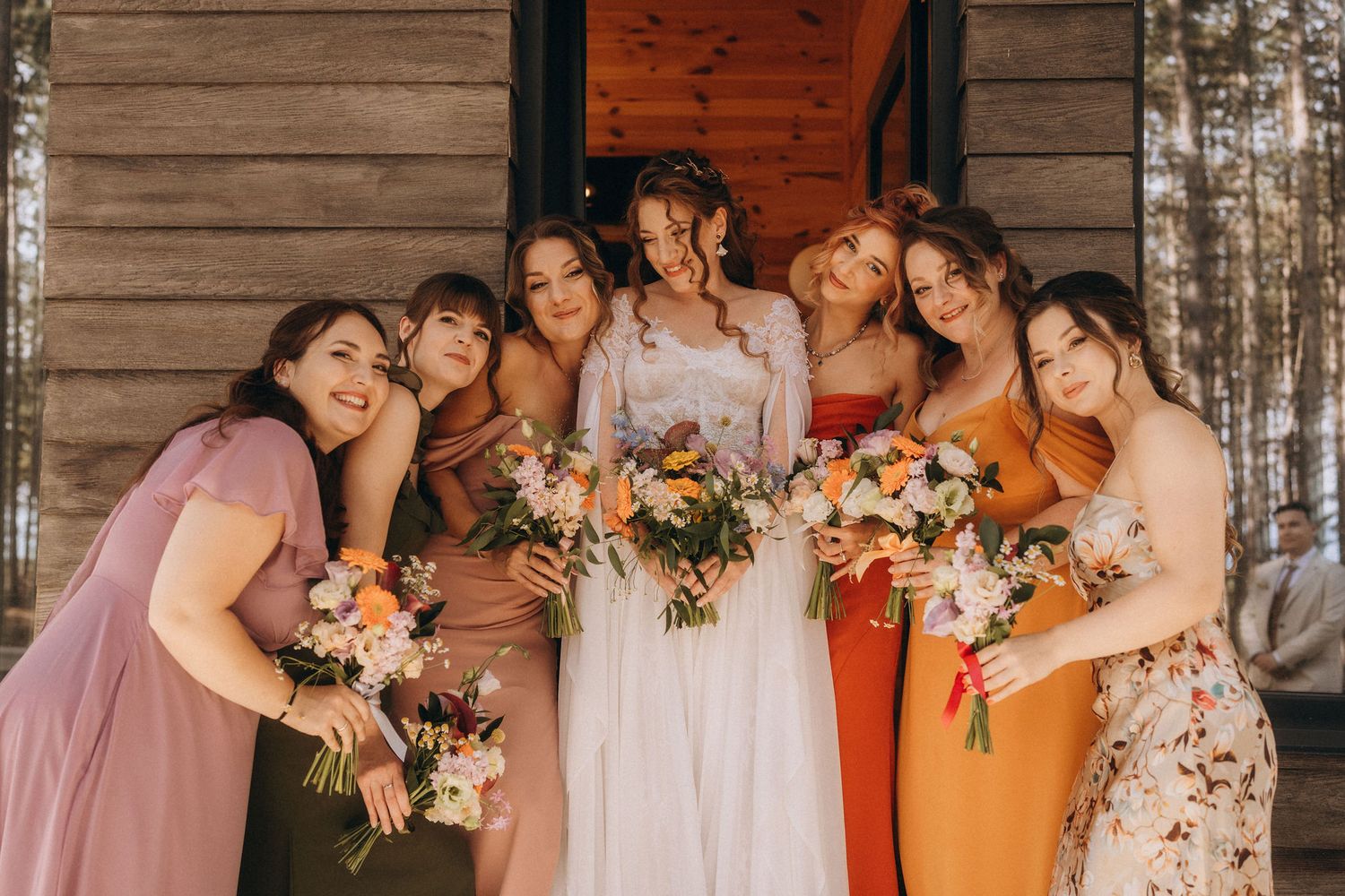 Bride in white gown poses with six bridesmaids in colorful dresses holding floral bouquets outside a rustic wooden cabin.