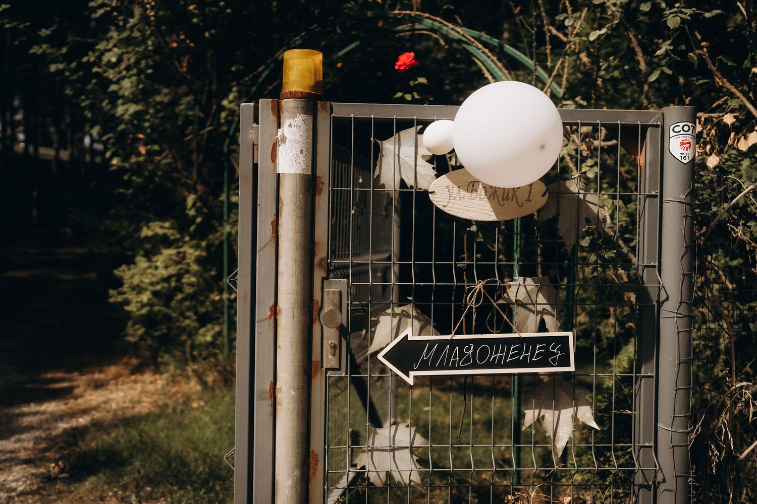 A metal gate decorated with white balloons and an arrow sign reading Mademoiselles surrounded by lush green trees.