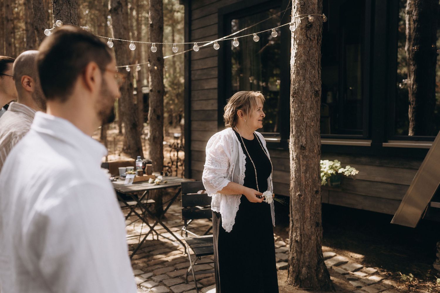 Outdoor wedding gathering with guests in formal attire socializing near a wooden cabin surrounded by tall trees.