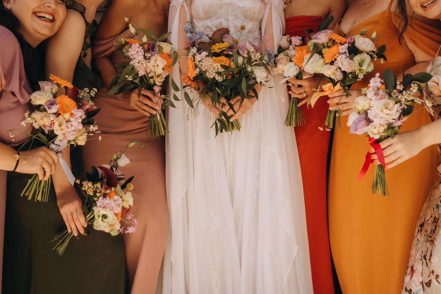 Bride in white gown with bridesmaids in colorful dresses holding vibrant floral bouquets at a wedding ceremony.