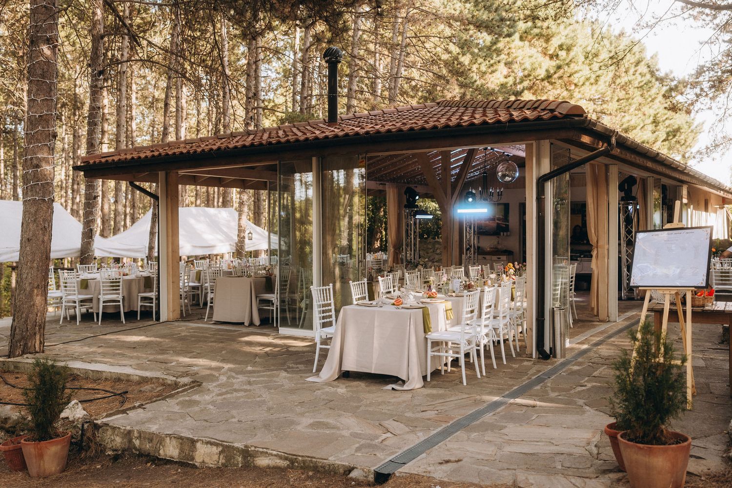 Elegant outdoor wedding venue with white chairs, draped tables, and a wooden pavilion surrounded by tall pine trees.