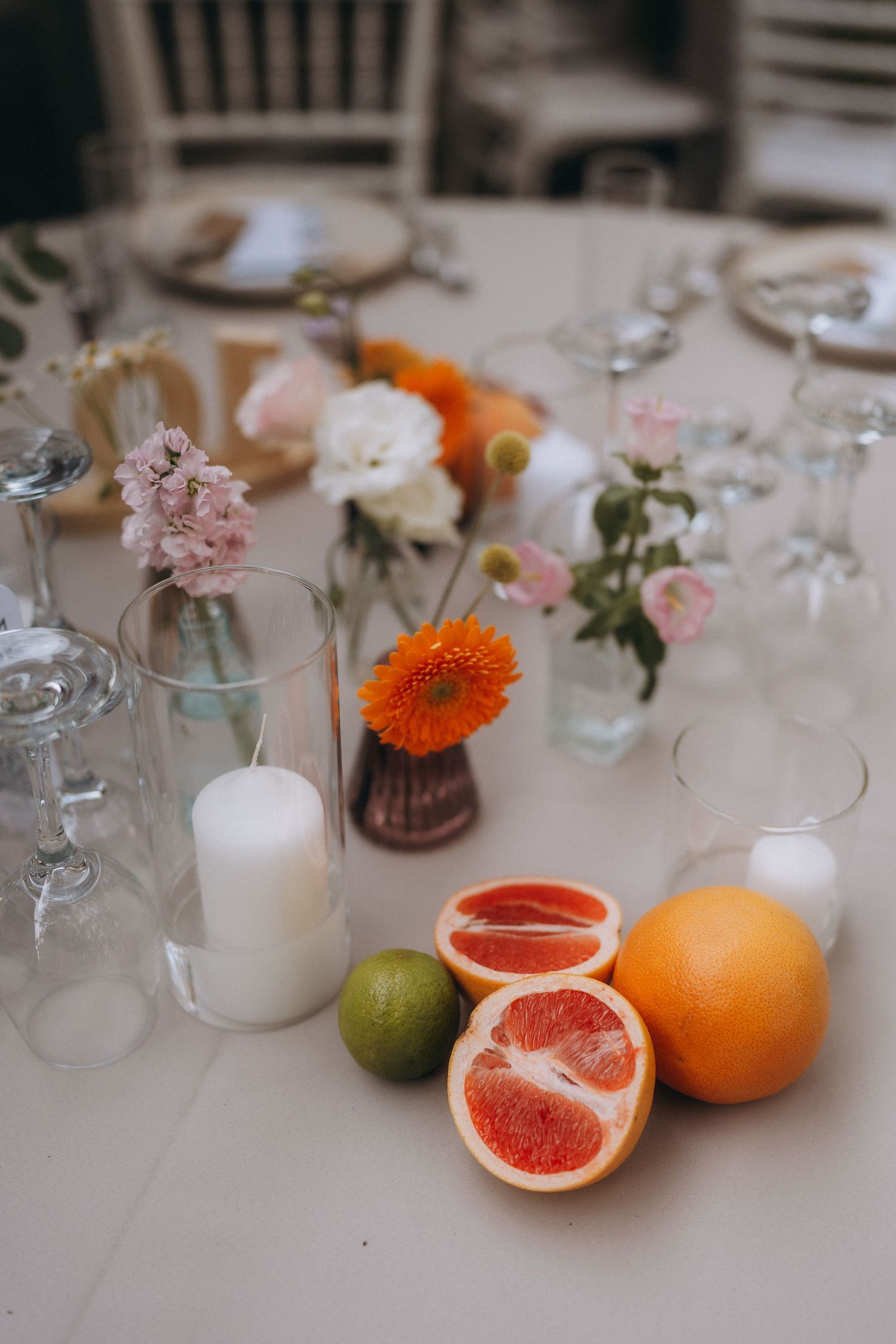 Elegant wedding table centerpiece with citrus fruits, white candles, and small floral arrangements in glass vases.