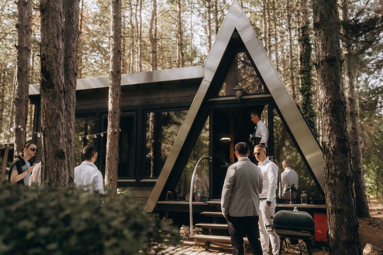 People gathered outside a modern A-frame cabin in a pine forest, dressed in formal attire for an outdoor wedding event.