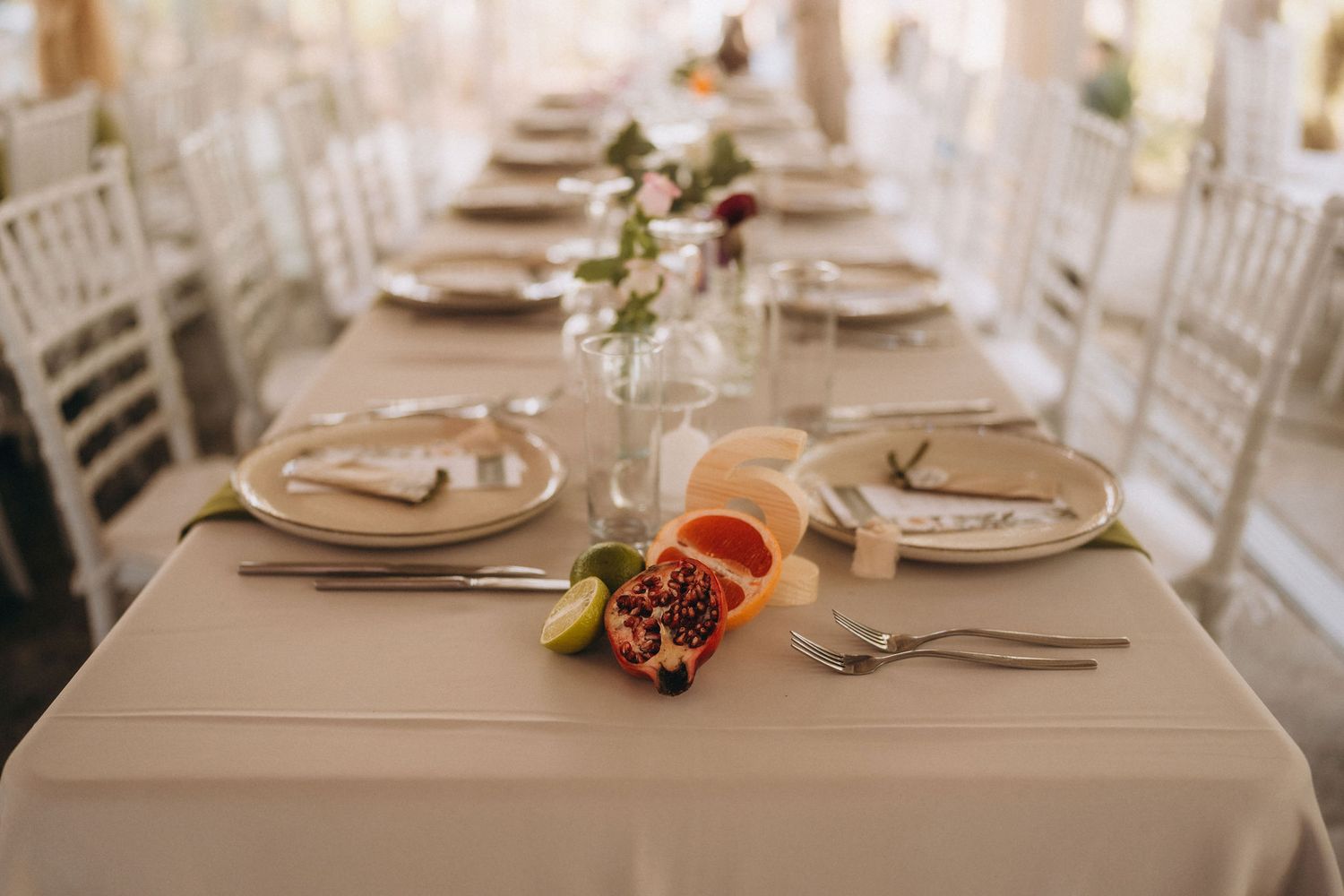 Elegant wedding reception table set with white plates, silverware, floral centerpieces, and fresh fruit garnishes on a cream tablecloth.