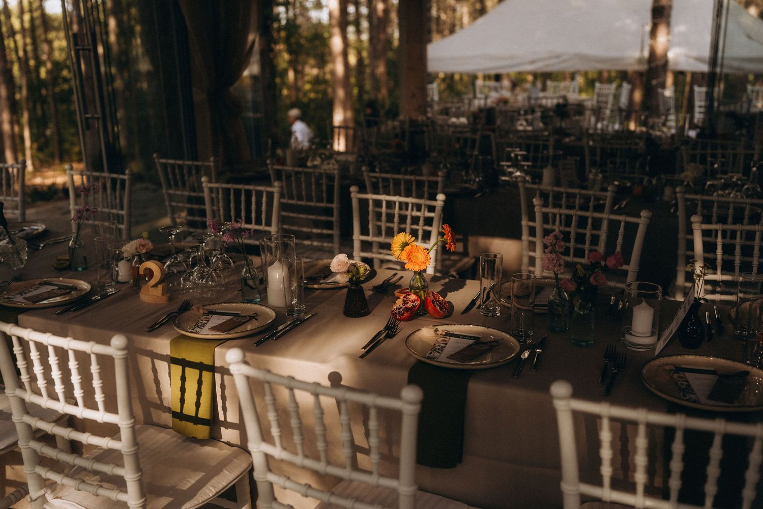 Elegant outdoor wedding reception tables with white chiavari chairs set among trees, featuring floral centerpieces and dark charger plates.