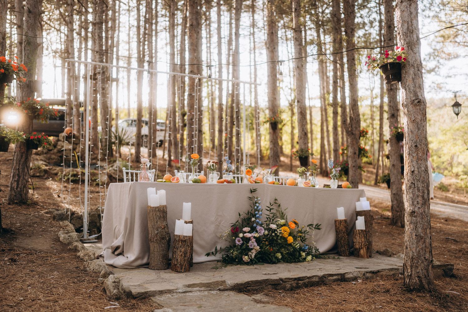 Elegant outdoor wedding reception table set in a sunlit pine forest with floral centerpieces and rustic log accents.