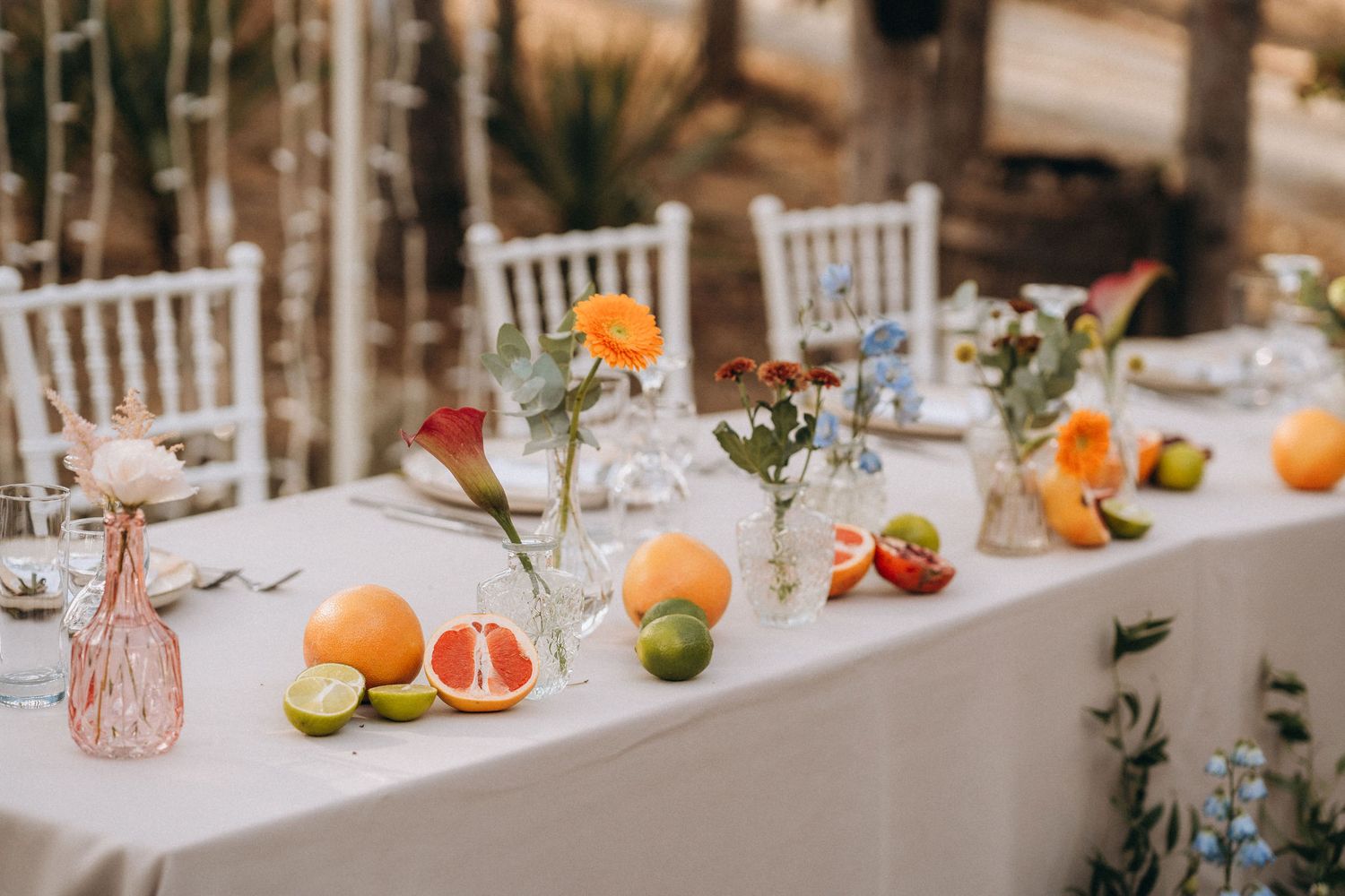 Elegant wedding table decorated with fresh citrus fruits, colorful flowers in glass vases, and white linen in an outdoor setting.