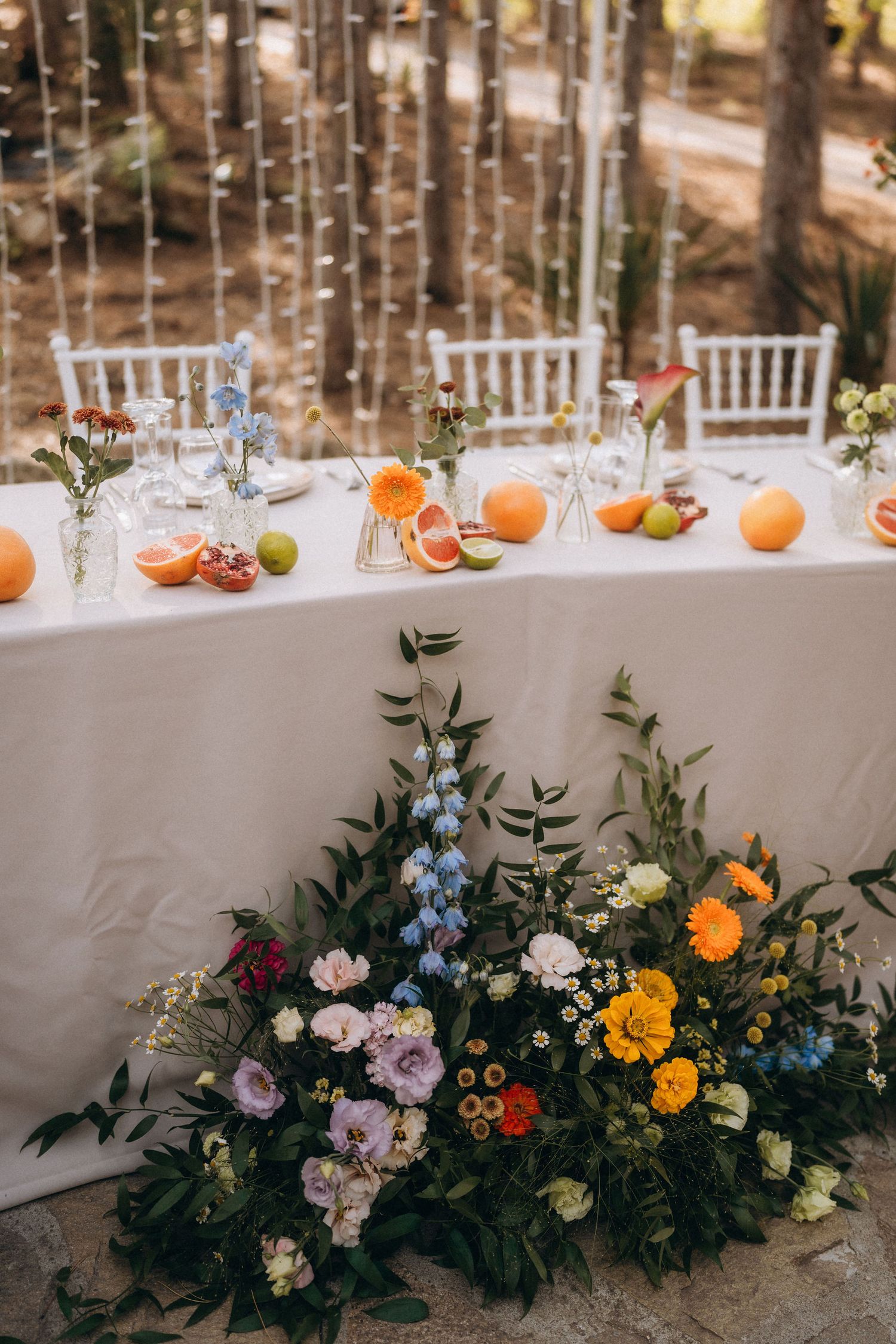Elegant outdoor wedding table with white linen, colorful flowers, and fresh citrus fruits, surrounded by trees.