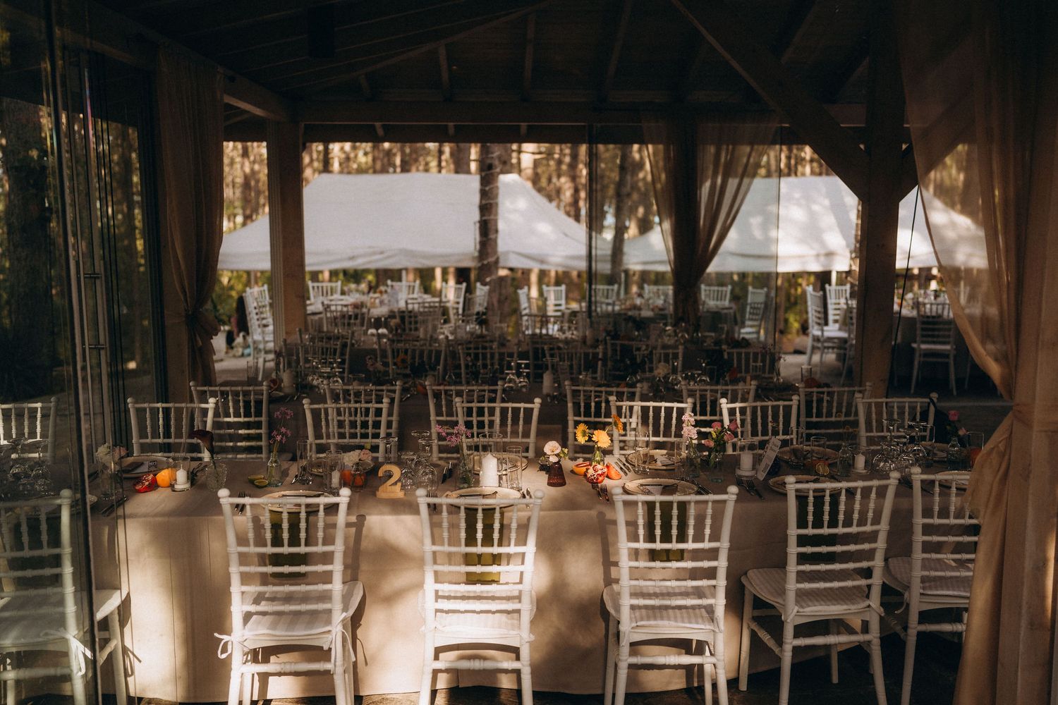 Elegant wedding reception venue with white Chiavari chairs, long draped tables, floral centerpieces, and a rustic open-air pavilion.