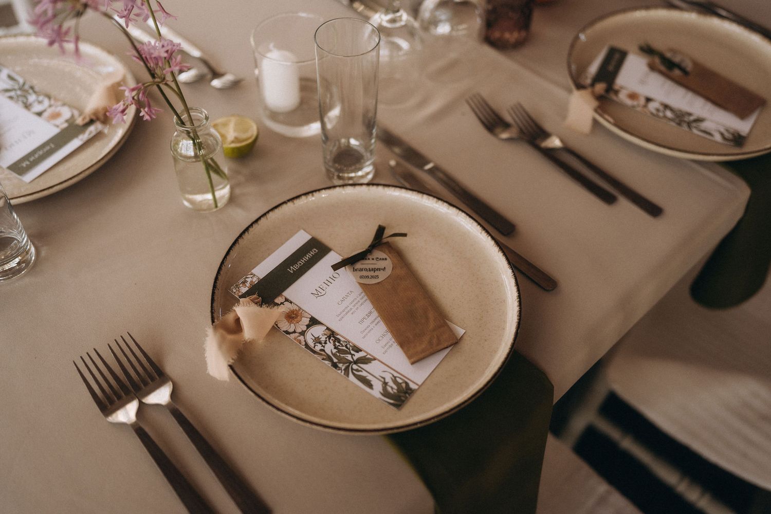 Elegant wedding table setting with cream plates, brown gift wrap, floral accents, candles, and silver cutlery on a beige tablecloth.