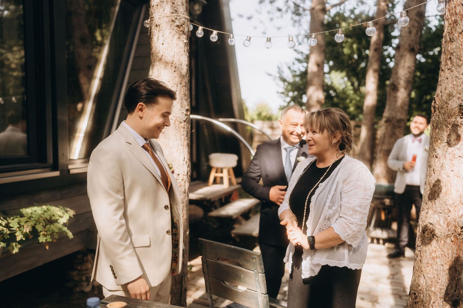 Groom in cream suit smiling with guests at an outdoor wedding ceremony surrounded by trees and string lights.