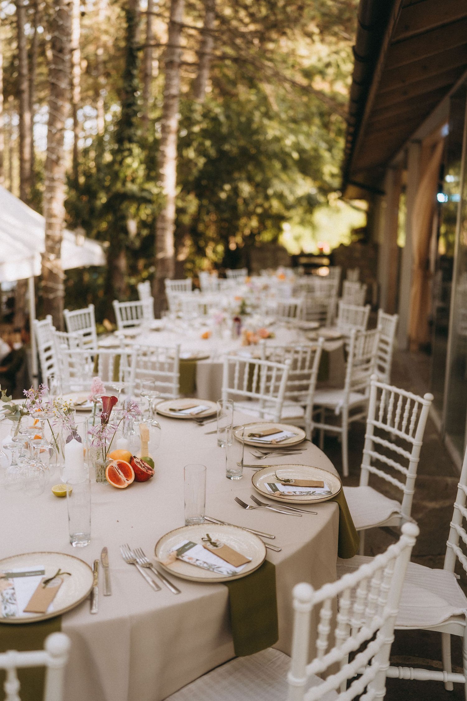 Elegant outdoor wedding reception with white Chiavari chairs, round tables, olive green napkins, and citrus centerpieces amid lush trees.