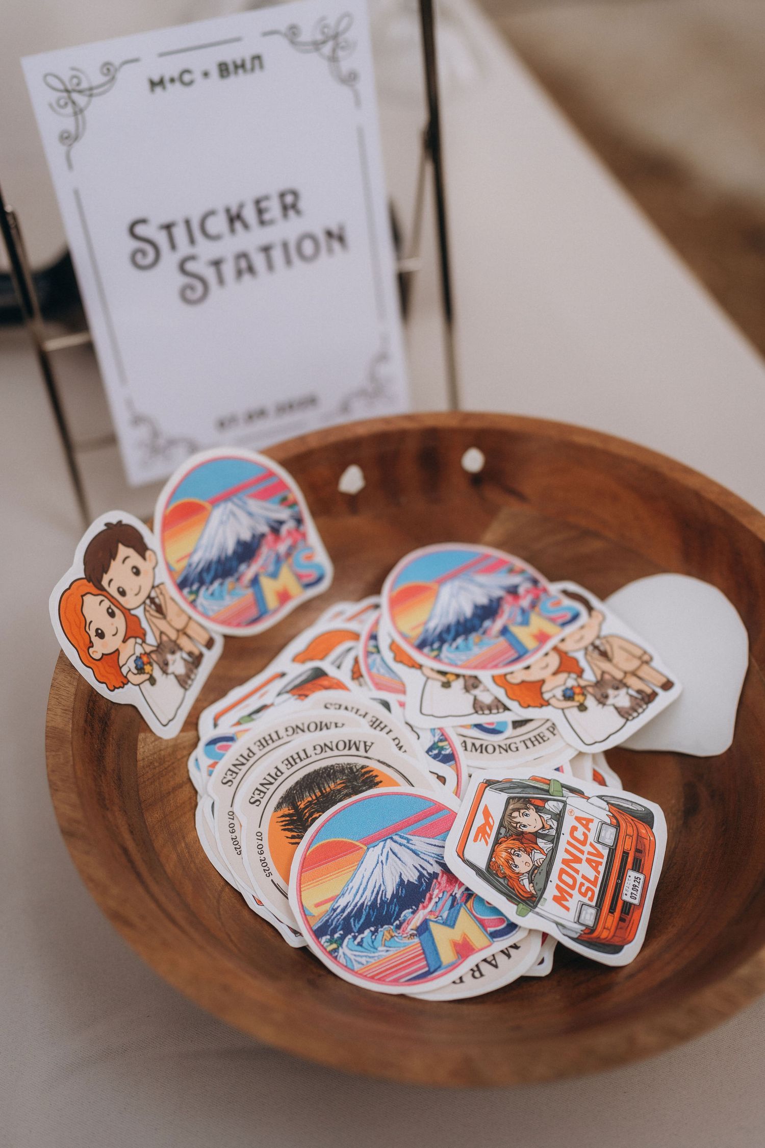 A wooden bowl filled with colorful travel-themed stickers near a Sticker Station sign at a wedding reception.