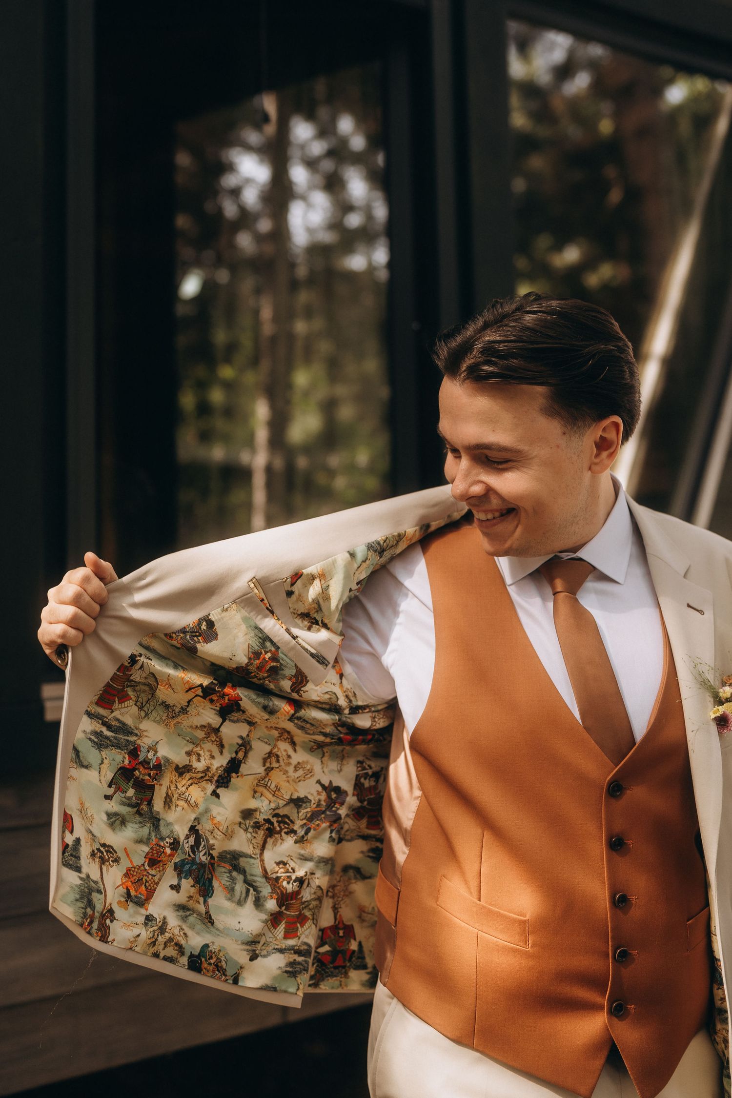 Smiling groom in beige suit with floral lining, orange vest and tie, showing jacket interior at wedding.