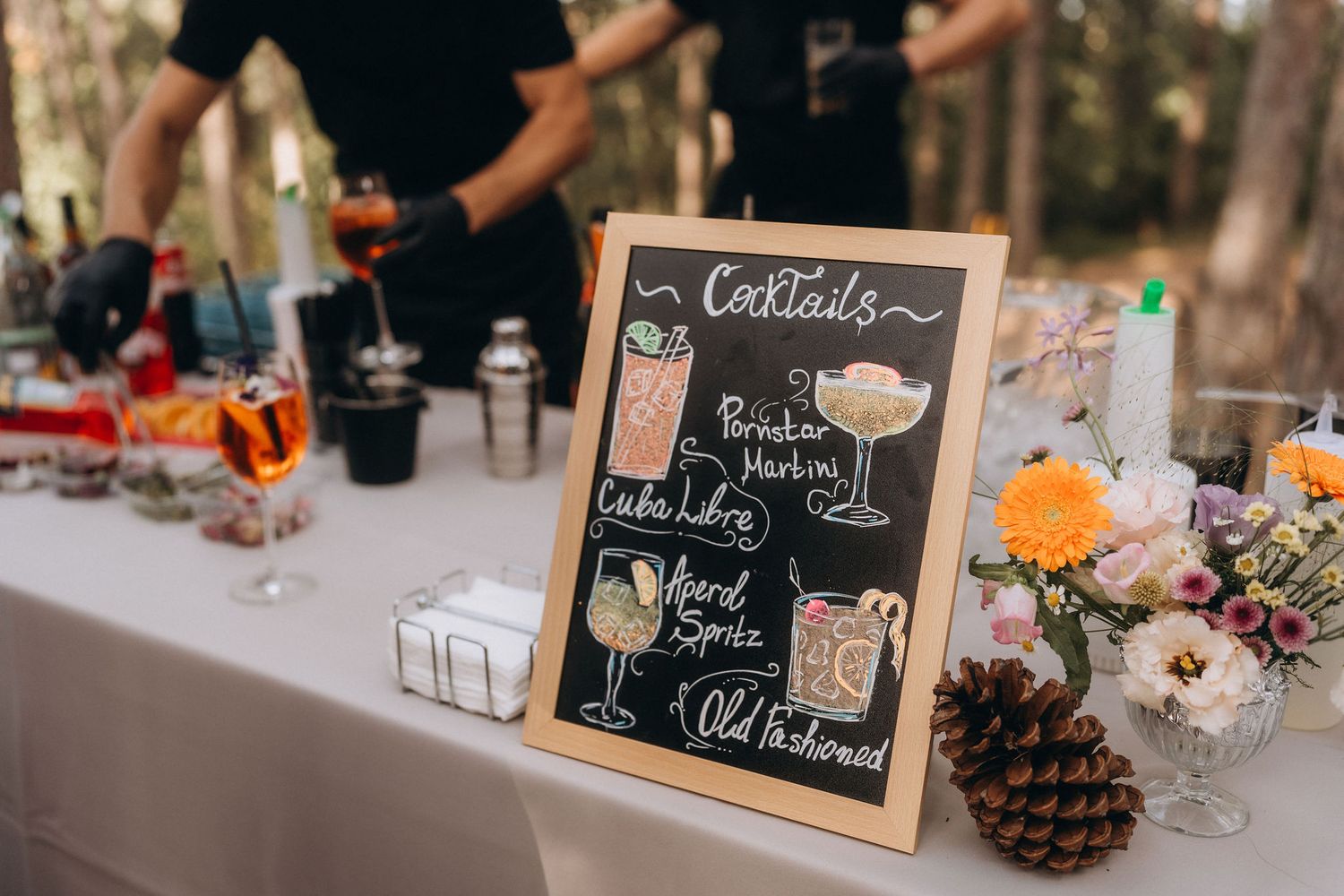 Chalkboard cocktail menu sign at outdoor wedding bar displaying drink options including Aperol Spritz and Old Fashioned.