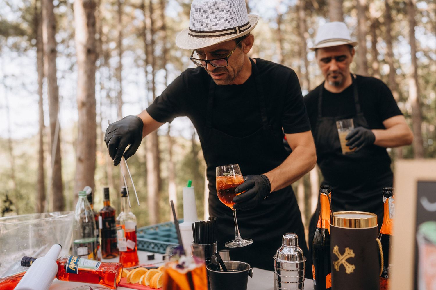 Two bartenders in black uniforms and white hats craft cocktails outdoors at a forest bar setup with bottles and shakers.