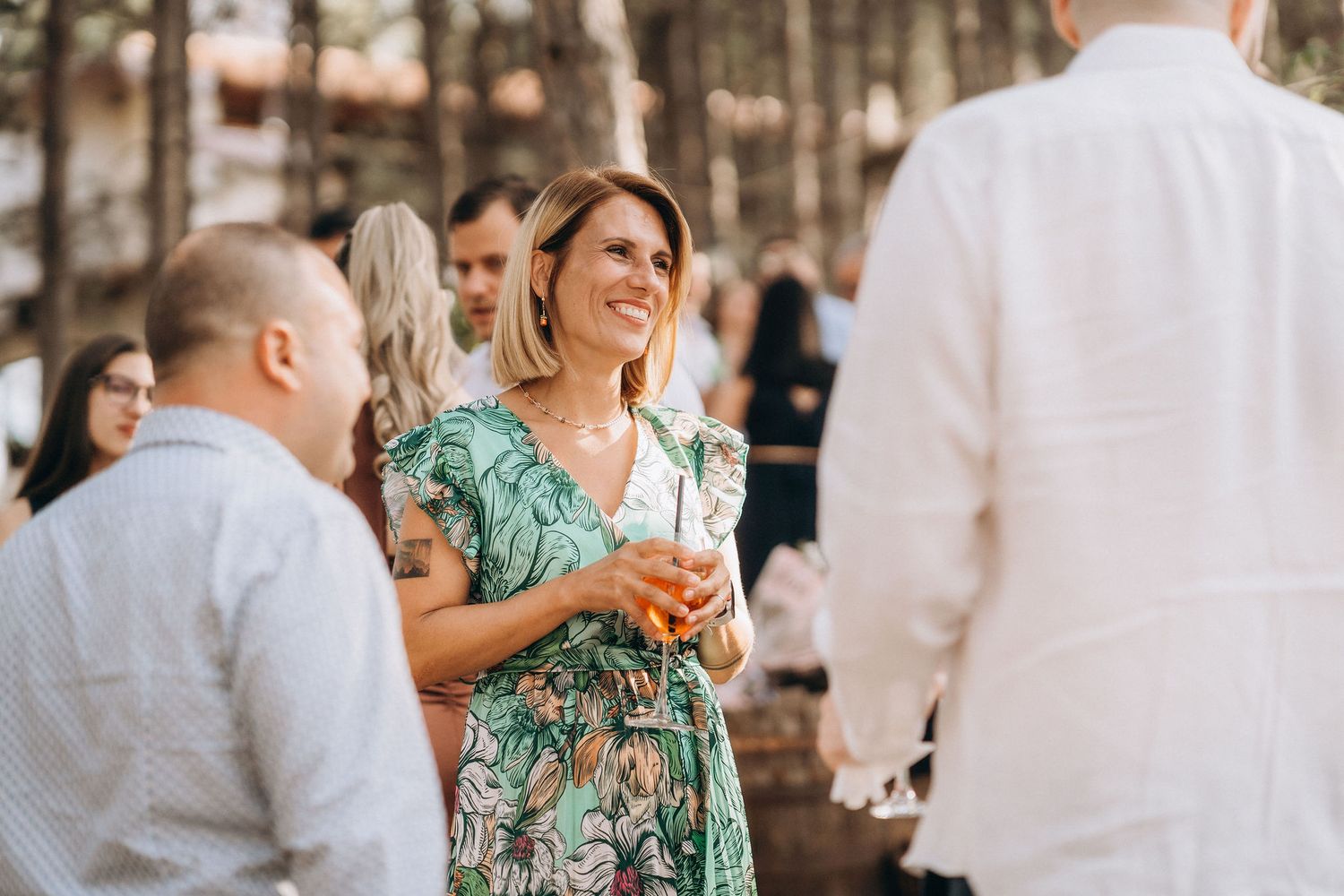 Woman in floral green dress smiling and holding a drink at an outdoor social gathering.