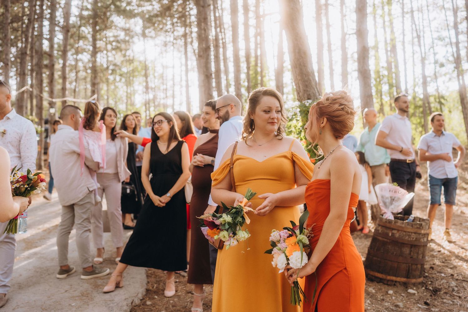 Bridesmaids in yellow and orange dresses holding bouquets at an outdoor forest wedding ceremony.