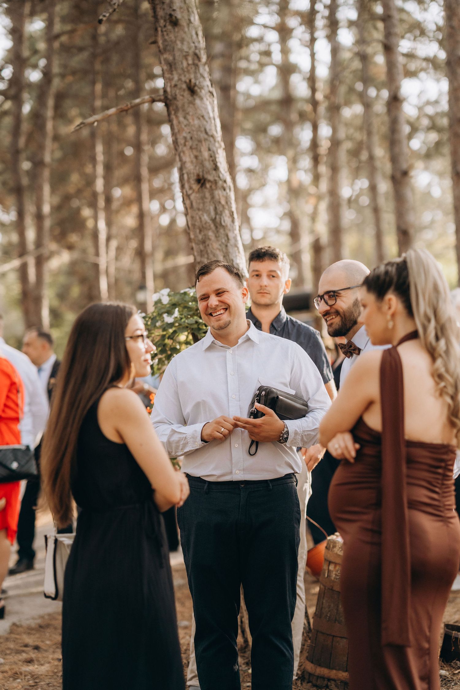 Guests socializing outdoors at a forest wedding, smiling man in white shirt holding camera among elegantly dressed attendees.