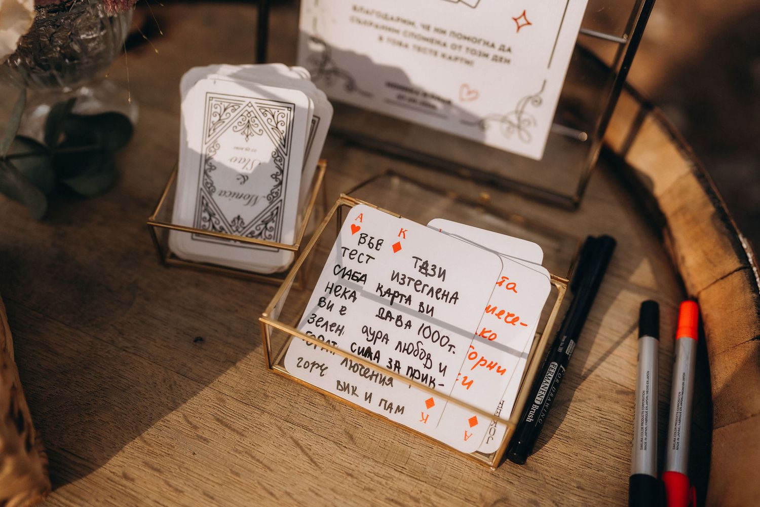 Close-up of handwritten notes in a gold box beside markers and playing cards on a wooden table.