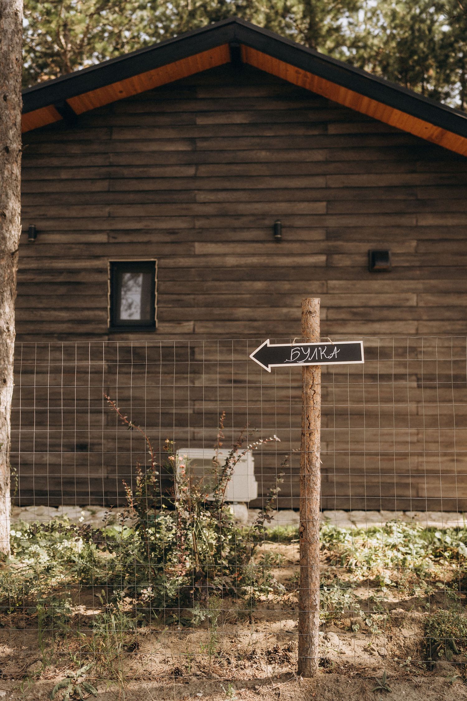 Rustic wooden cabin with a directional arrow sign reading Булка mounted on a post in front of a wire fence.