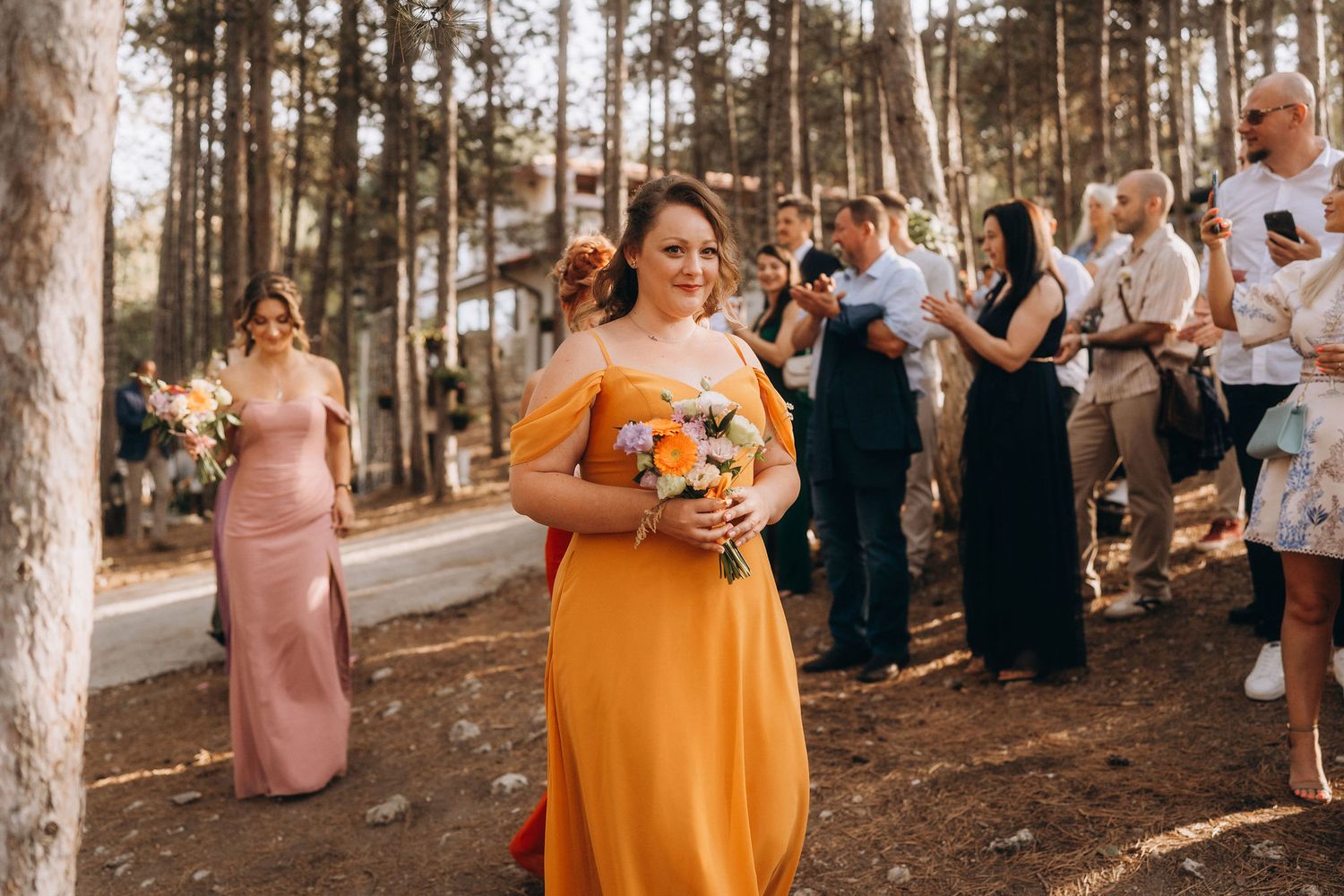 Bridesmaid in orange off-shoulder gown holding bouquet walks down outdoor aisle at forest wedding ceremony.