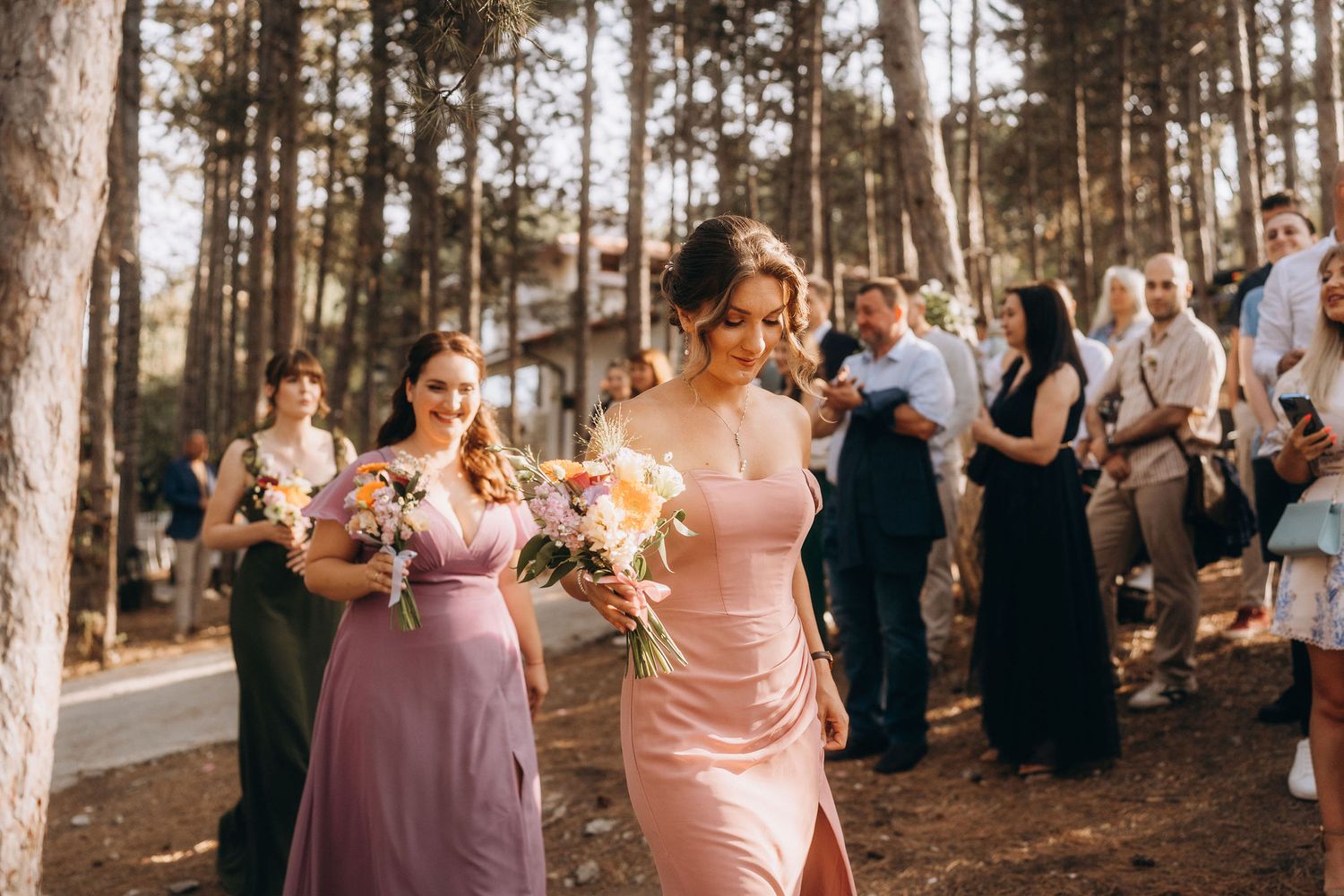 Bridesmaids in pink and mauve gowns walk through a sunlit forest wedding ceremony holding colorful bouquets.