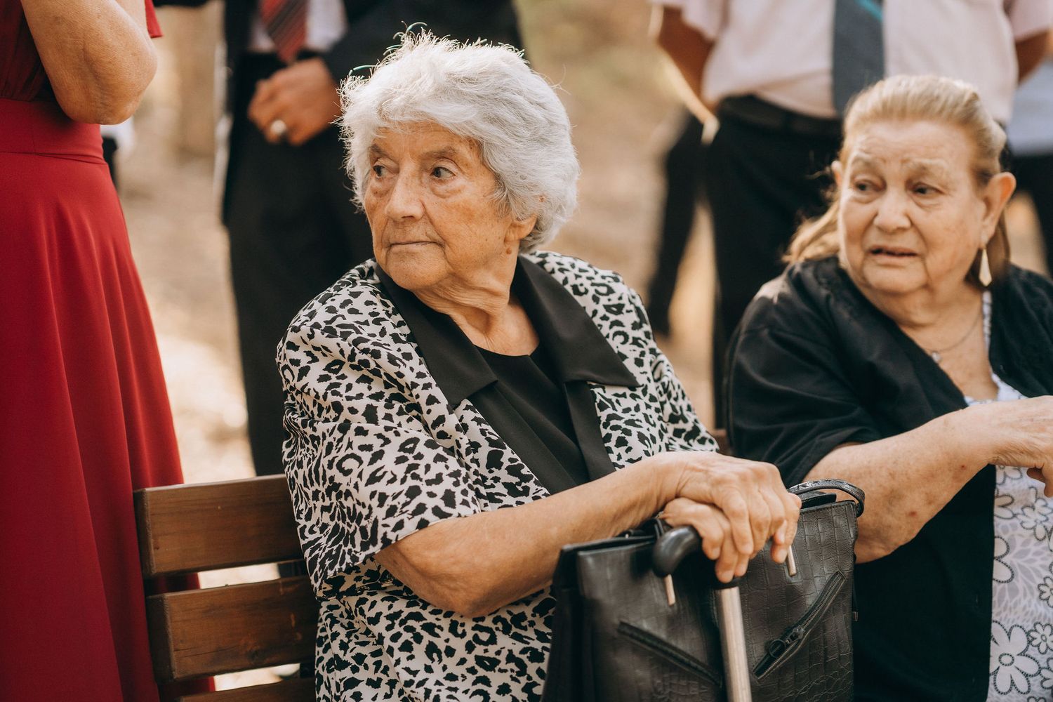 Two elderly women sitting outdoors, one holding a cane, dressed in black at a social gathering.