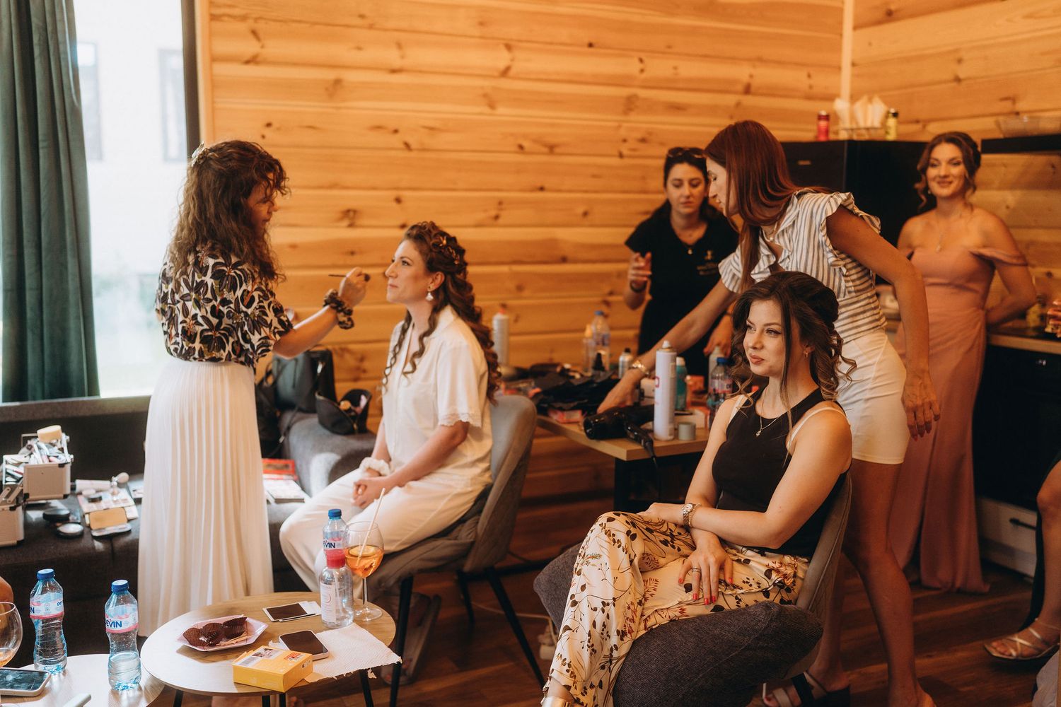 Women getting hair and makeup done in a rustic wooden cabin during a bridal party preparation session.