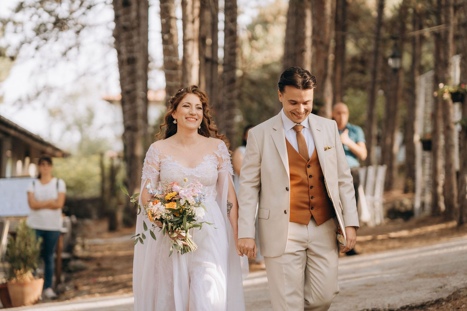 Bride and groom walking hand in hand down an outdoor aisle surrounded by pine trees on their wedding day.