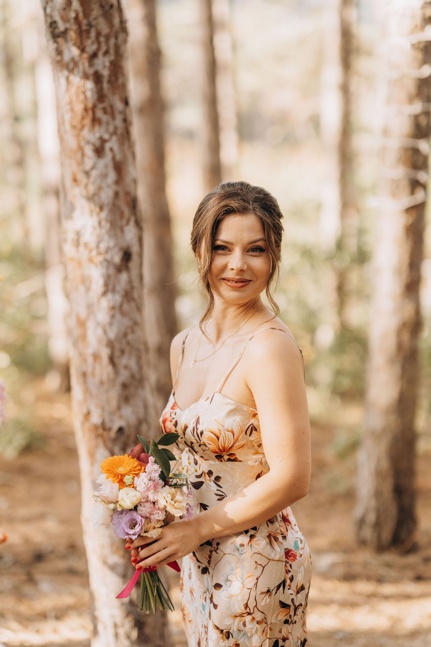 Woman in floral dress holding colorful bouquet smiling in sunlit pine forest.