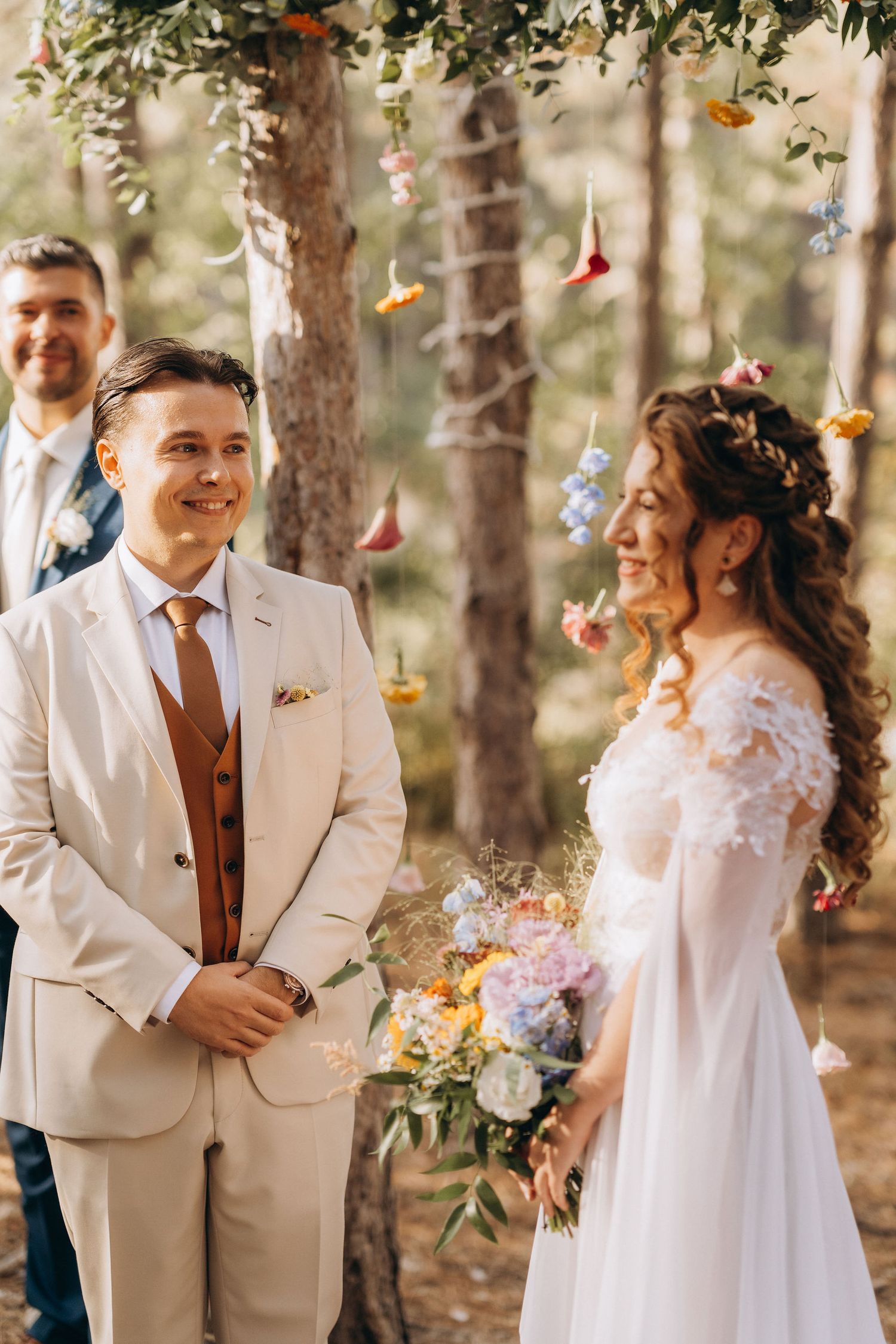 Smiling groom in beige suit with brown vest stands facing bride in white lace dress during outdoor forest wedding ceremony.