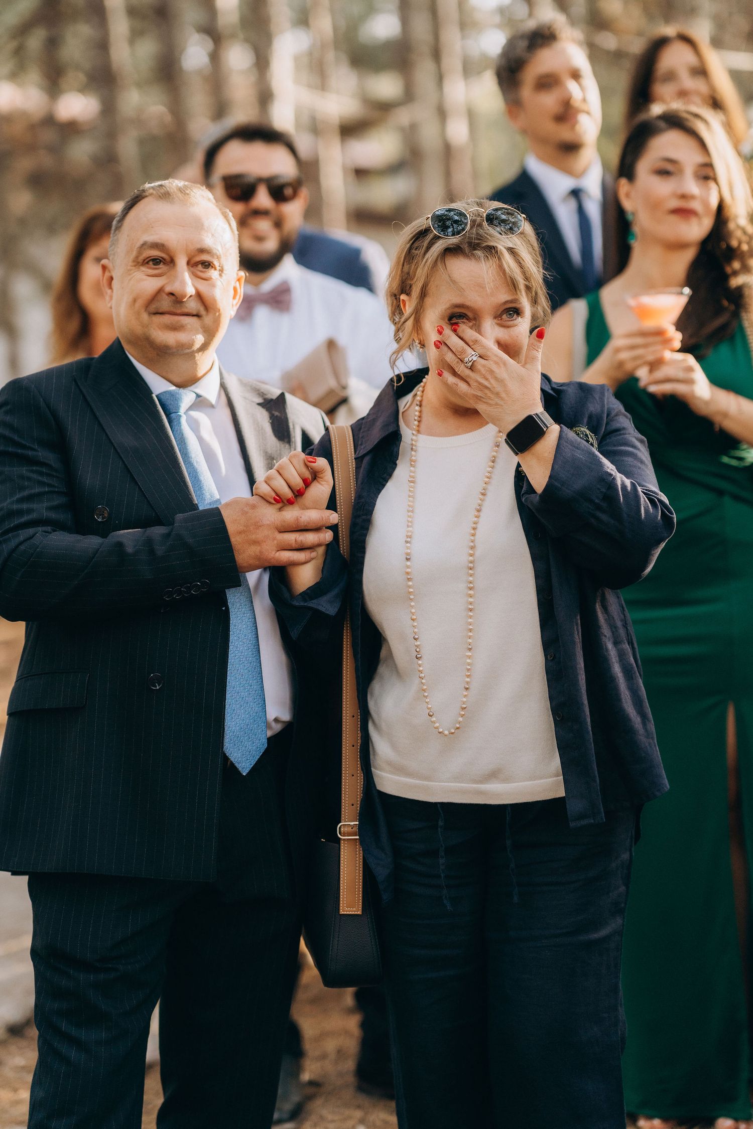 Emotional wedding guests react with joy, woman covering mouth in surprise, man in navy suit beside her, group celebrating outdoors.