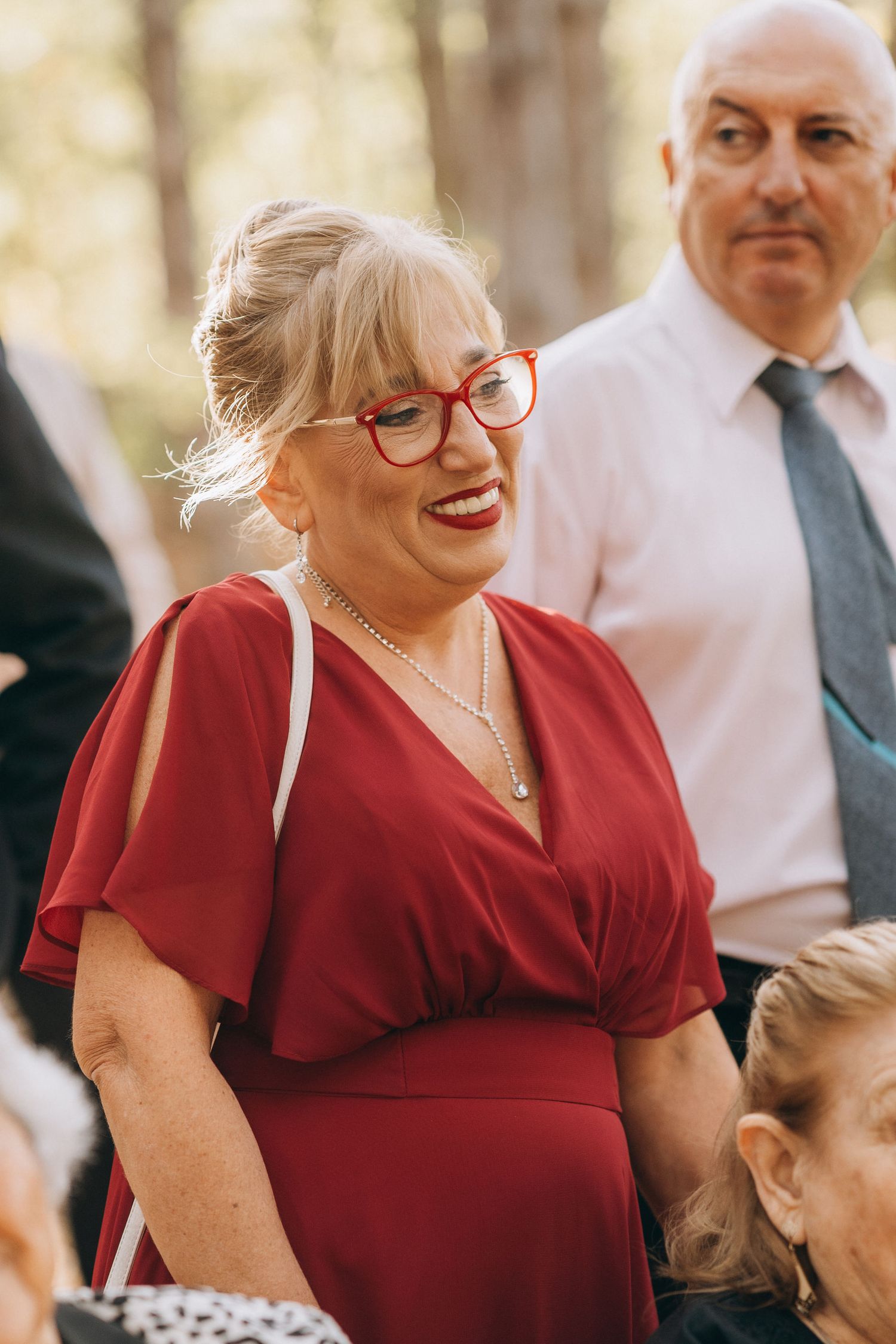 Smiling blonde woman in red dress and red glasses at outdoor event with man in white shirt and tie behind her.