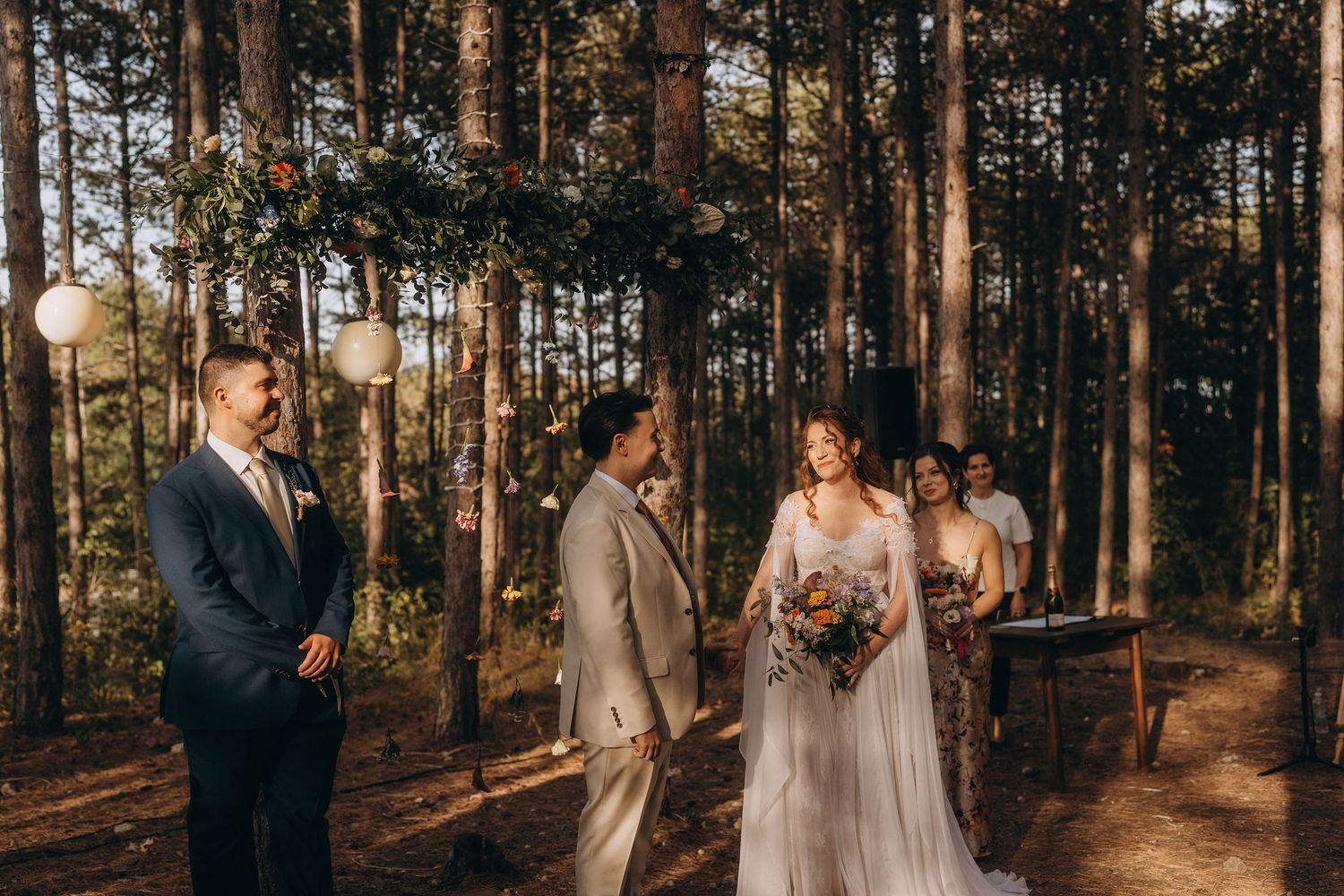 Outdoor forest wedding ceremony with bride in white gown and groom in navy suit standing among tall pine trees.