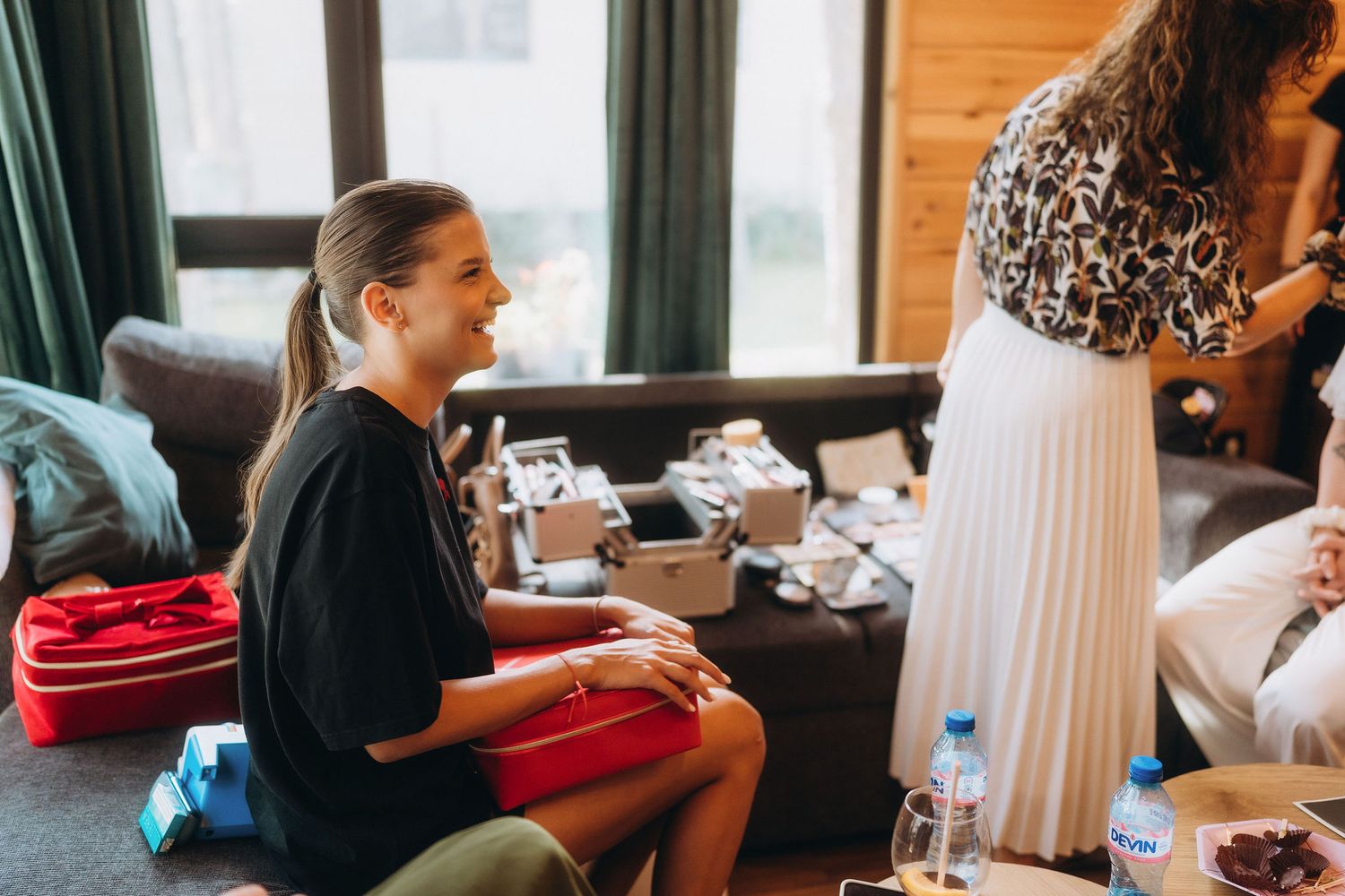 A smiling woman in a black dress sits on the floor during a bridal preparation session in a cozy wooden interior.