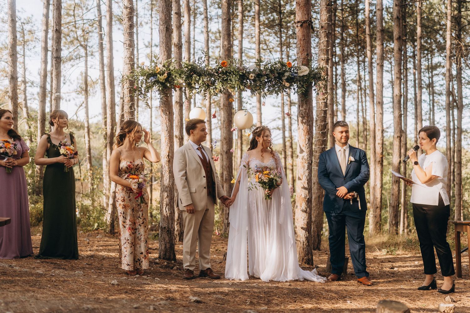 Outdoor wedding ceremony in a pine forest with bride, groom, officiant, bridesmaids, and groomsmen under a floral arch.
