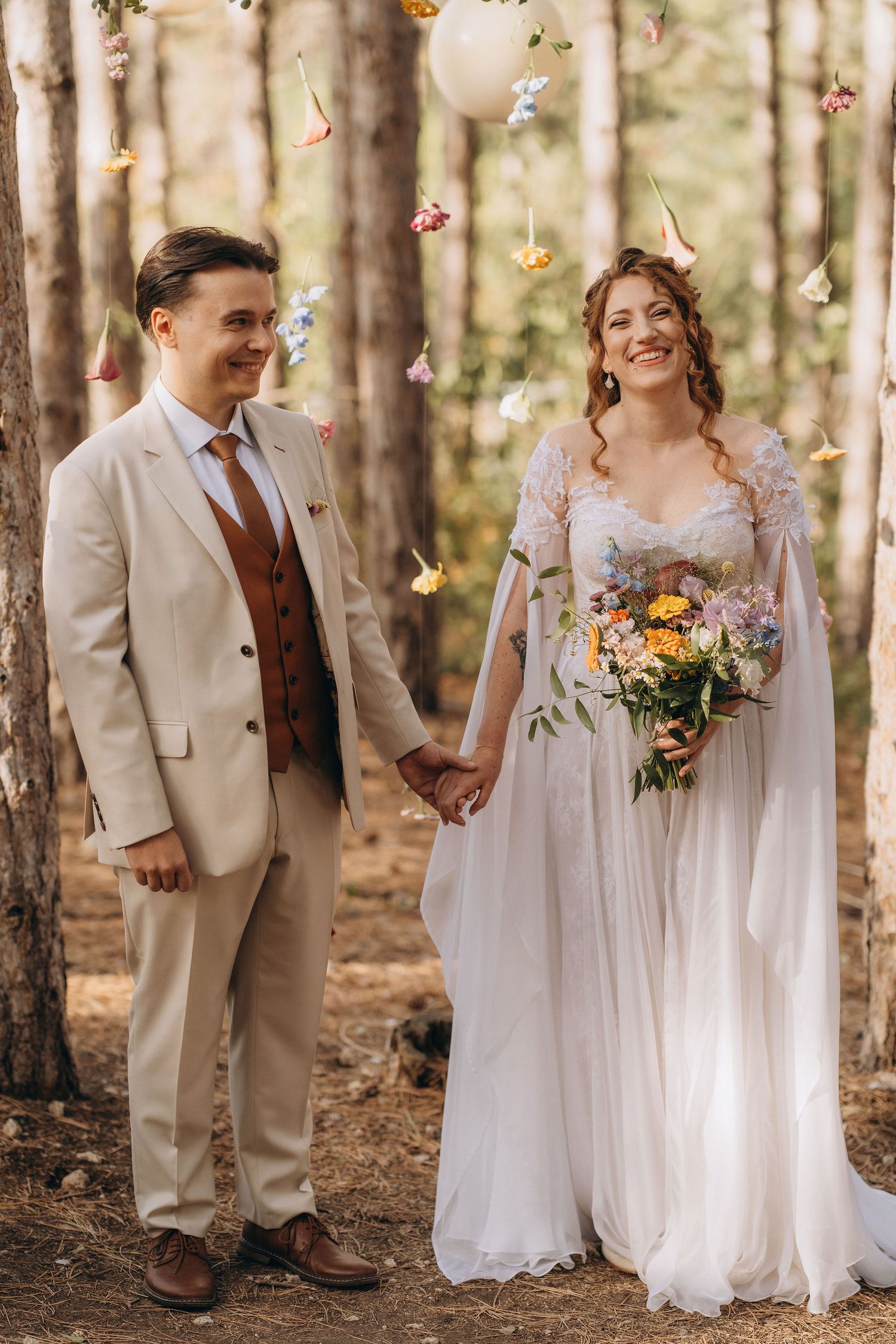 Bride and groom holding hands in a forest wedding ceremony with colorful hanging flowers.